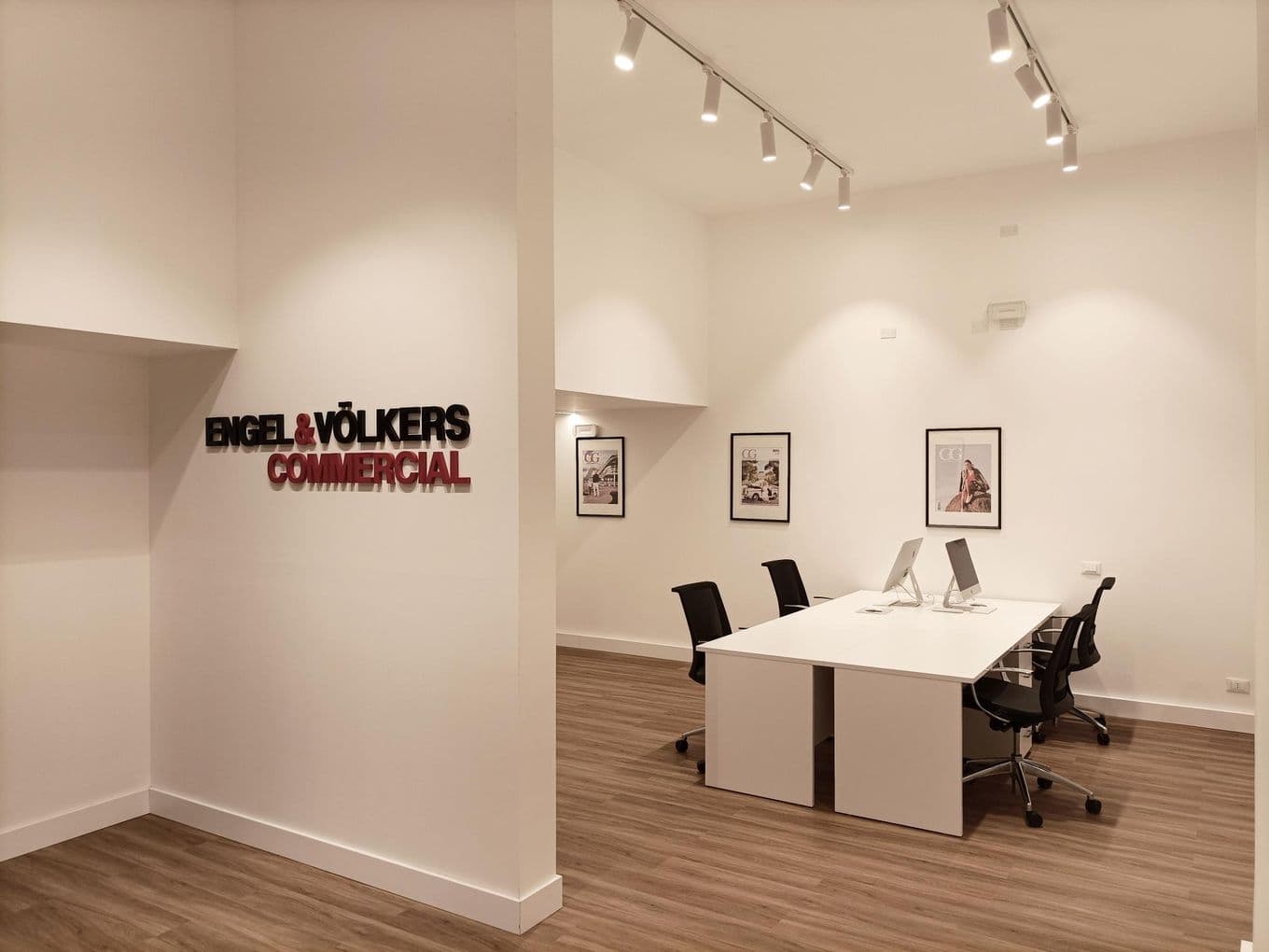 Modern office space with white walls, wooden floors, a sleek desk with computers, and framed pictures on the walls. Signage reads "ENGEL & VÖLKERS COMMERCIAL".