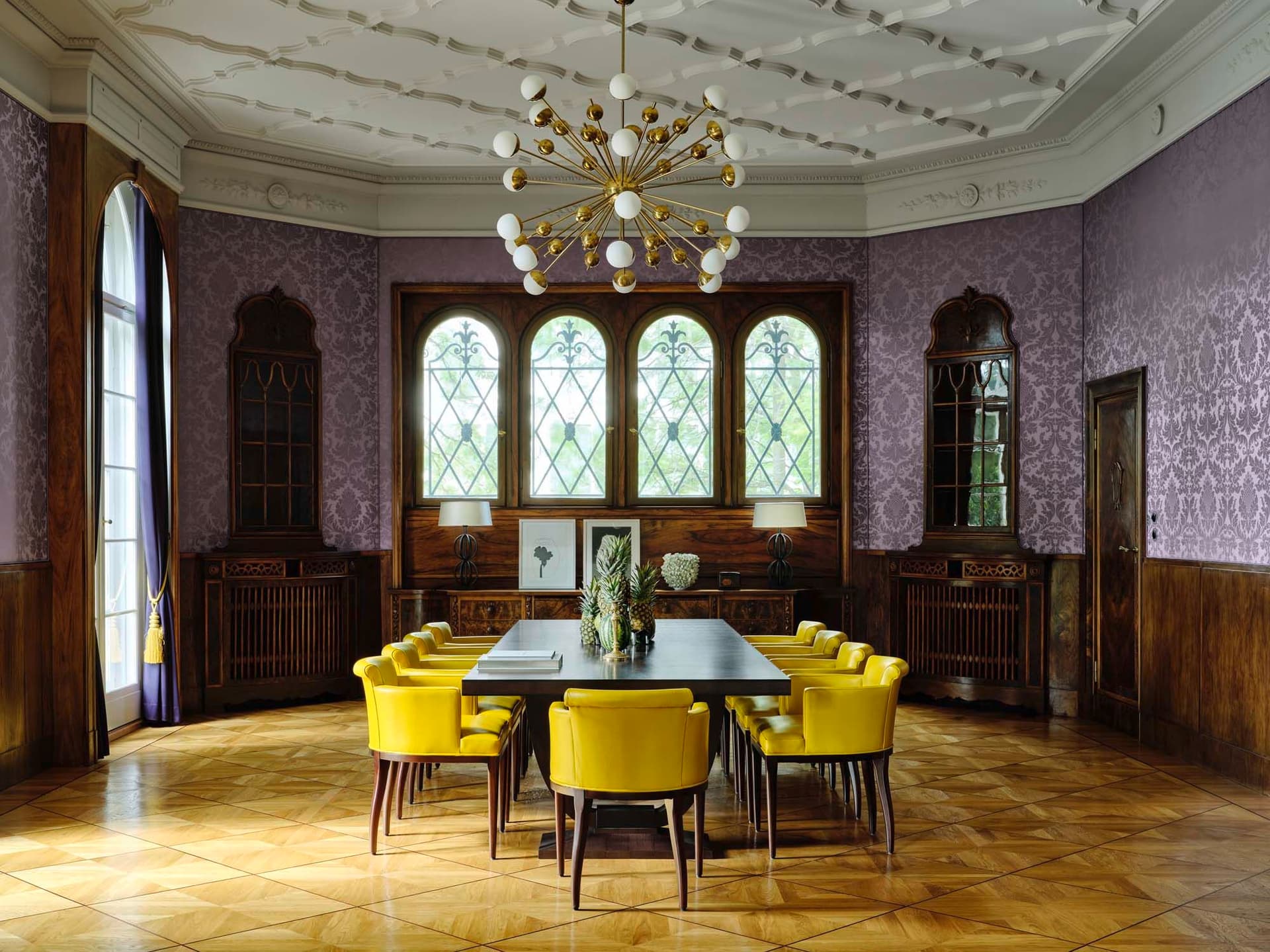 Opulent dining room with yellow chairs, a dark wood table, and a starburst chandelier. Walls are purple with damask pattern.