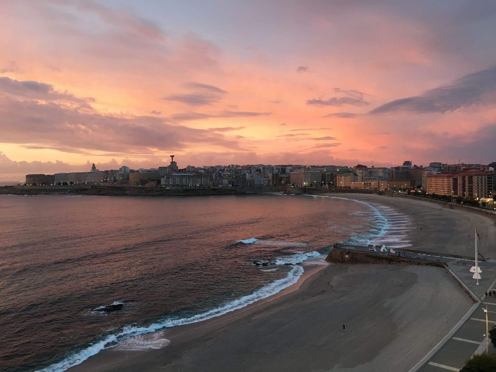 Ein idyllischer Sonnenuntergang mit pastellfarbenem Himmel in Rosa- und Lilatönen über einer geschwungenen Bucht. Die sanften Wellen des Meeres treffen auf den Sandstrand, während eine Stadt mit zahlreichen Gebäuden und Straßenlichtern im Hintergrund liegt. Eine Promenade verläuft entlang des Strandes, und vereinzelte Spaziergänger genießen die ruhige Atmosphäre.