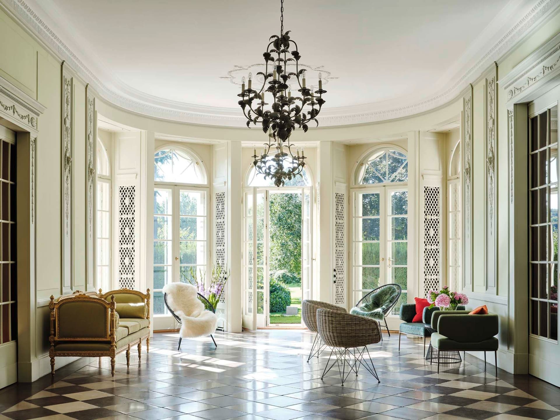 Bright sunroom with black and white checkered floors, ornate chandelier, and various chairs. Large windows open to a green garden.