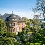 Aerial view of a salmon-colored mansion with a dark roof, surrounded by lush green gardens and a river in the background.