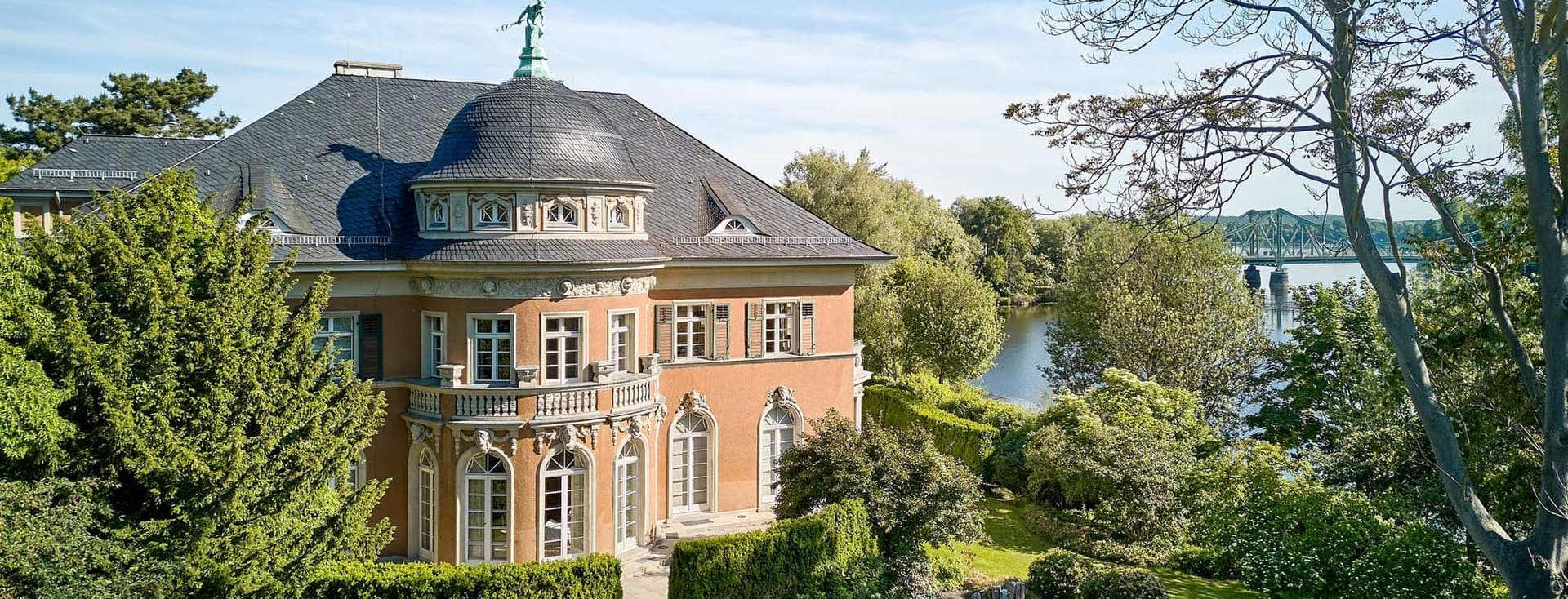 Aerial view of a salmon-colored mansion with a dark roof, surrounded by lush green gardens and a river in the background.