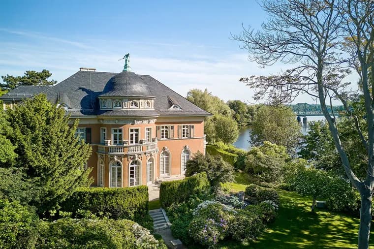Aerial view of a salmon-colored mansion with a dark roof, surrounded by lush green gardens and a river in the background.