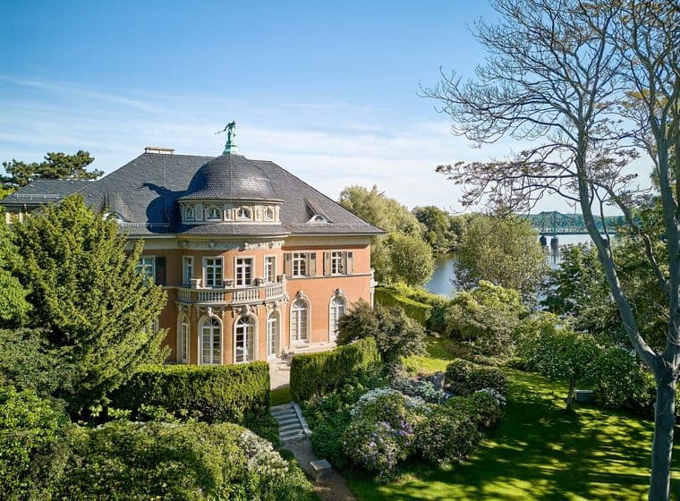 Aerial view of a salmon-colored mansion with a dark roof, surrounded by lush green gardens and a river in the background.