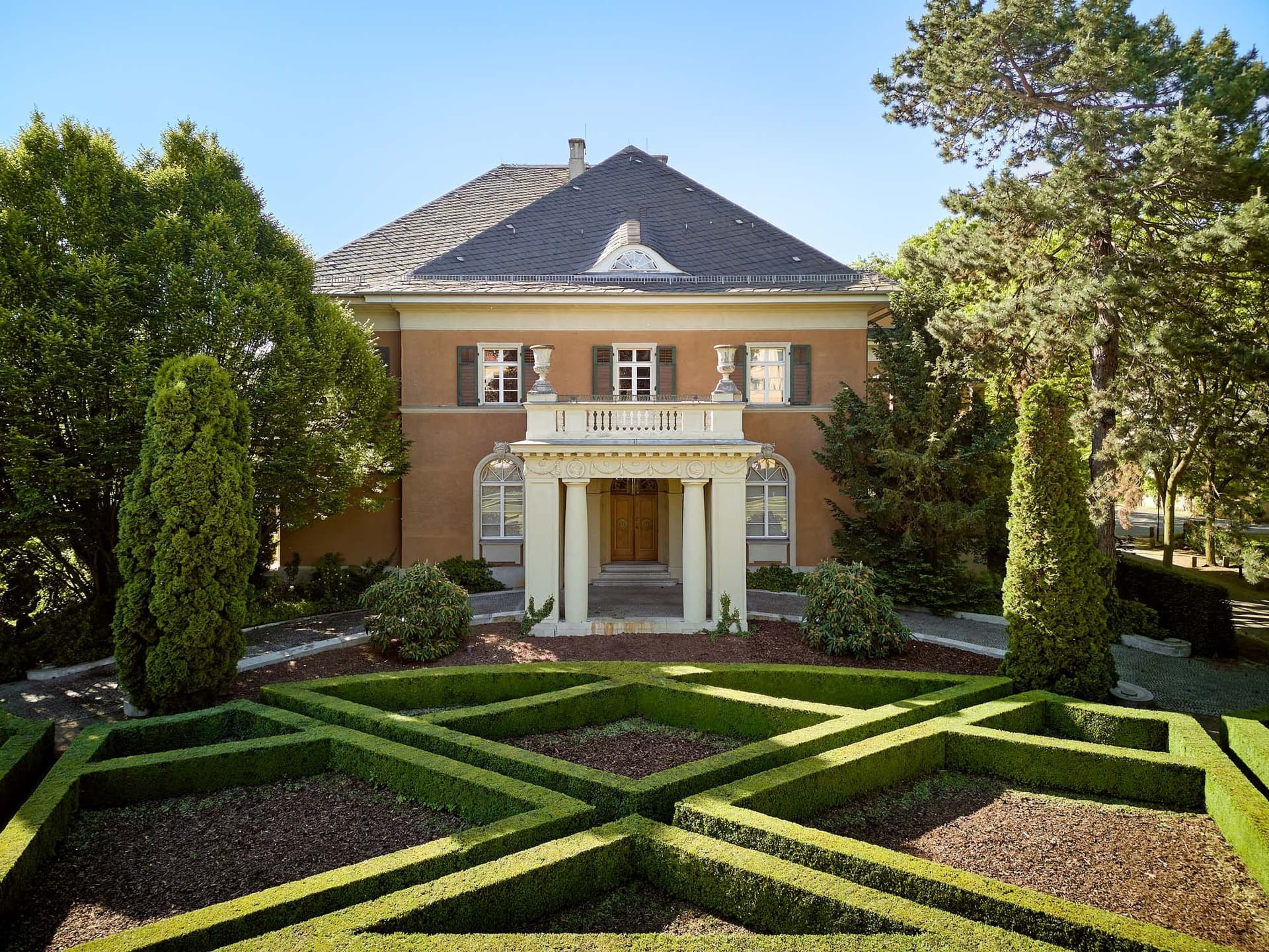 Two-story house with a gray roof, brown walls, and a white columned porch. A green hedge maze is in the front yard.