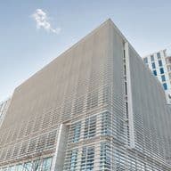 Moderne architektonische Außenansicht des Engel & Völkers-Hauptquartiers mit einer eleganten Fassade mit horizontaler Metallverkleidung, raumhohen Fenstern und einem unverwechselbaren kantigen Design vor einem hellblauen Himmel. Das angrenzende Wohnhochhaus ist im Hintergrund zu sehen.