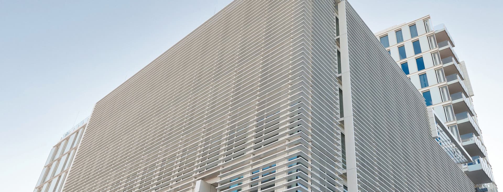 Modern architectural exterior view of the Engel & Völkers headquarters building featuring a sleek facade with horizontal metal cladding, floor-to-ceiling windows, and a distinctive angular design against a light blue sky. Adjacent high-rise residential building visible in the background.
