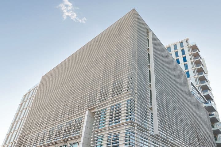 Modern architectural exterior view of the Engel & Völkers headquarters building featuring a sleek facade with horizontal metal cladding, floor-to-ceiling windows, and a distinctive angular design against a light blue sky. Adjacent high-rise residential building visible in the background.