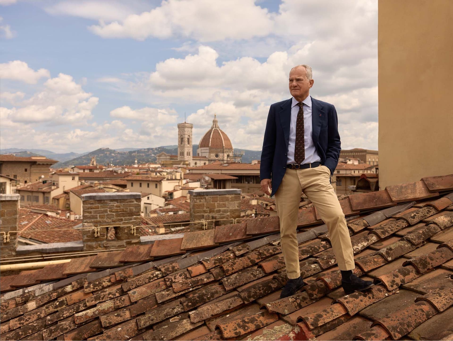 A man in a suit stands confidently on a rooftop terrace overlooking Florence, with the Cathedral of Santa Maria del Fiore in the background.