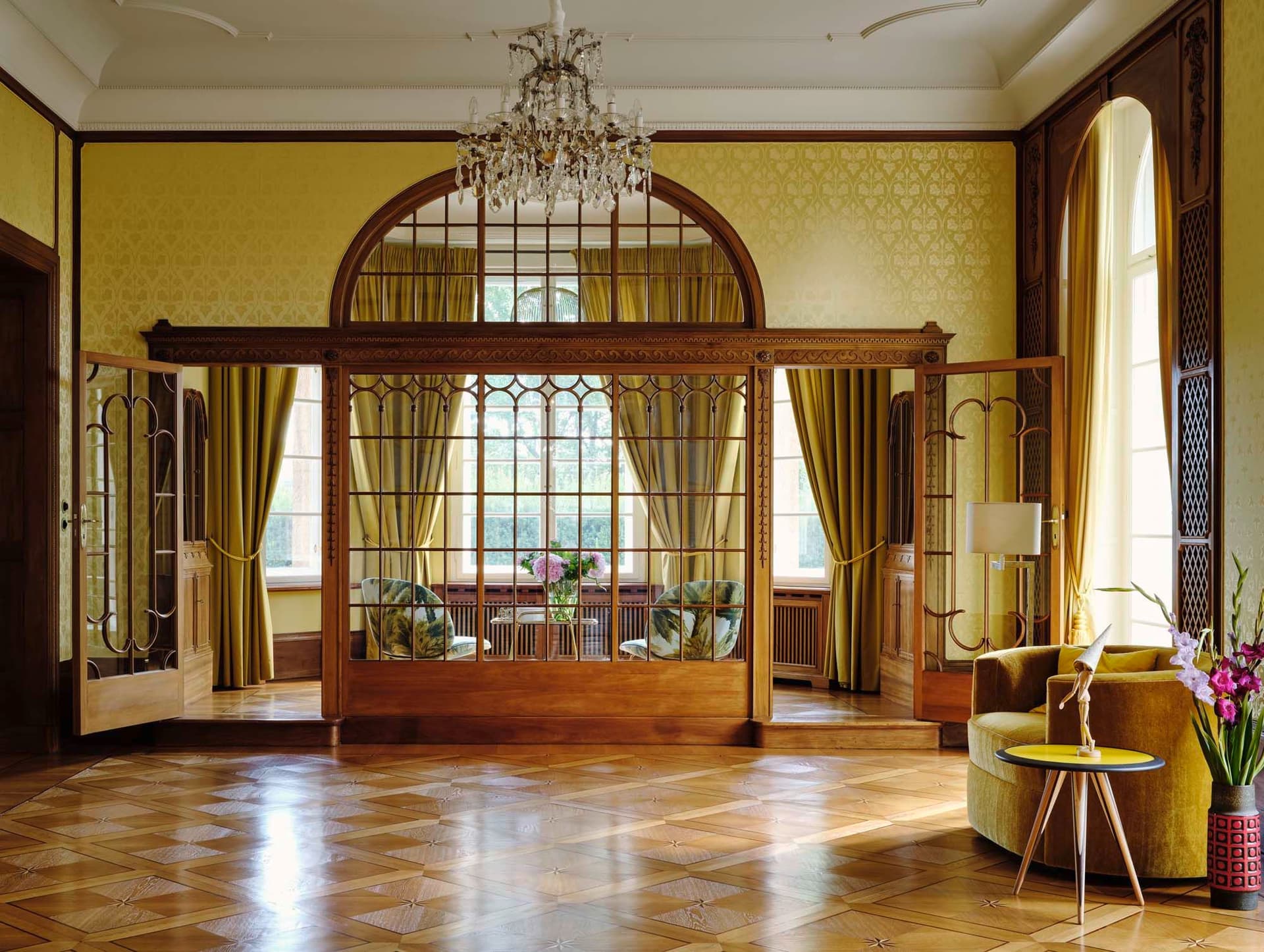 Elegant room with parquet floor, yellow walls, and wood-framed glass doors. A chandelier hangs above, and a seating area is visible through the doors.