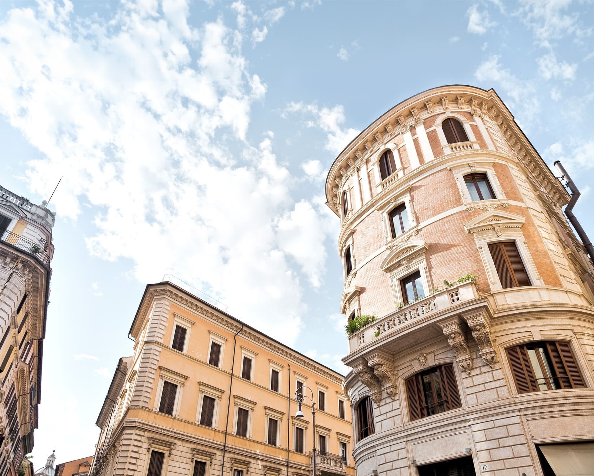 A view of magnificent historic buildings in central Rome on a sunny day. The foreground is dominated by an impressive ochre-colored corner building with ornate decorations and balconies, exemplifying typical Roman architecture. Adjacent are other classic city houses. A blue sky dotted with white clouds provides a picturesque backdrop. This striking urban scene illustrates the allure and high value of real estate in the Italian capital, reflecting the steep property prices in Rome's historic center. The image captures the timeless beauty that contributes to the city's enduring real estate appeal