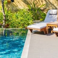 Due sedie a sdraio in legno con cuscini bianchi sono posizionate accanto a una piscina blu, con piante verdi e una parete ruvida sullo sfondo.