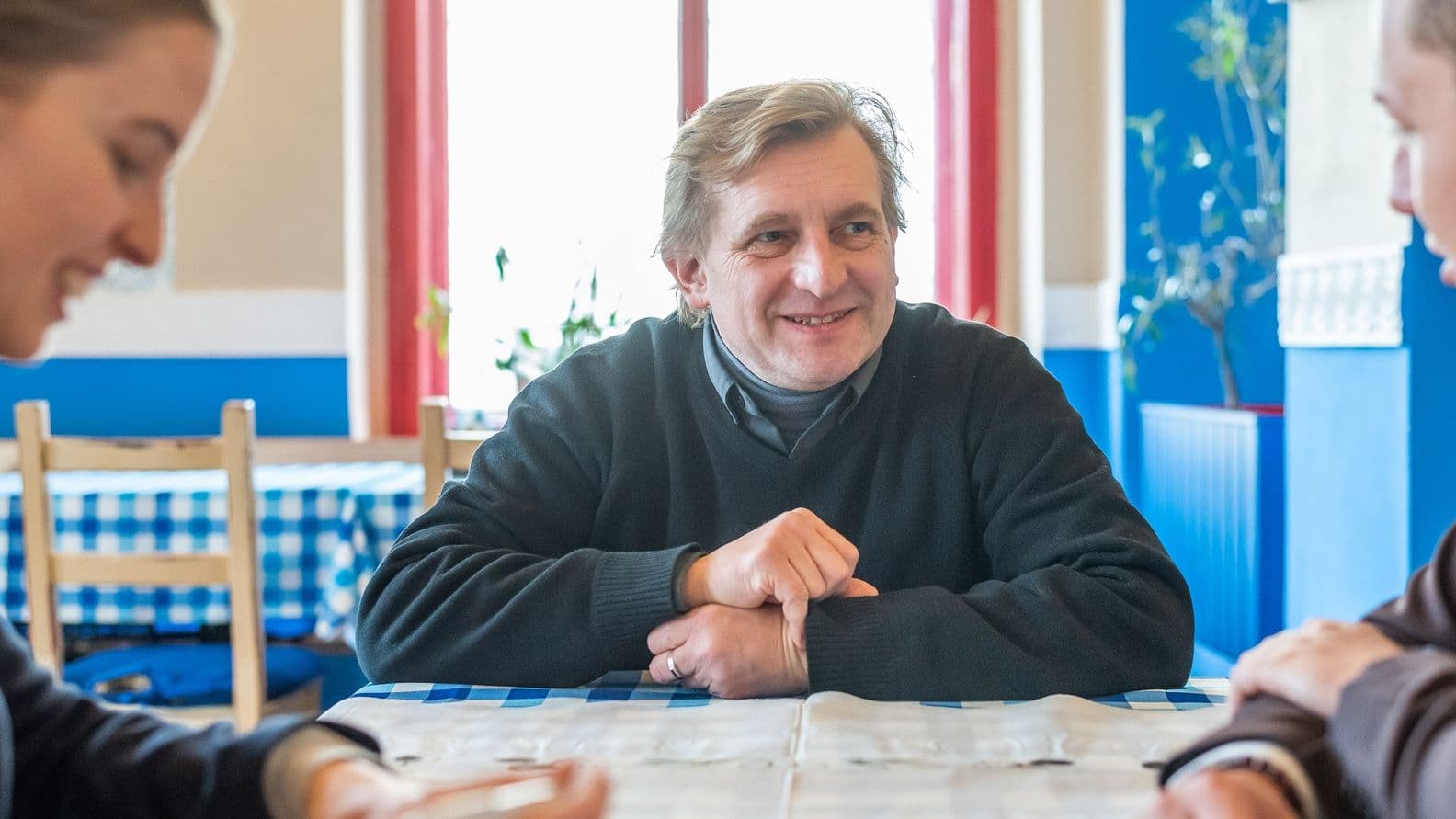 Man in a sweater sitting at a table in a blue and white interior, smiling and talking to people off-frame. Window and plants in the background.