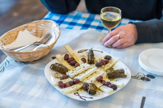 Plate with Greek specialties – cheese, olives, and stuffed vine leaves – on a table with a blue-and-white cloth. Next to it, a glass of white wine and a person’s hand in a dark sweater.