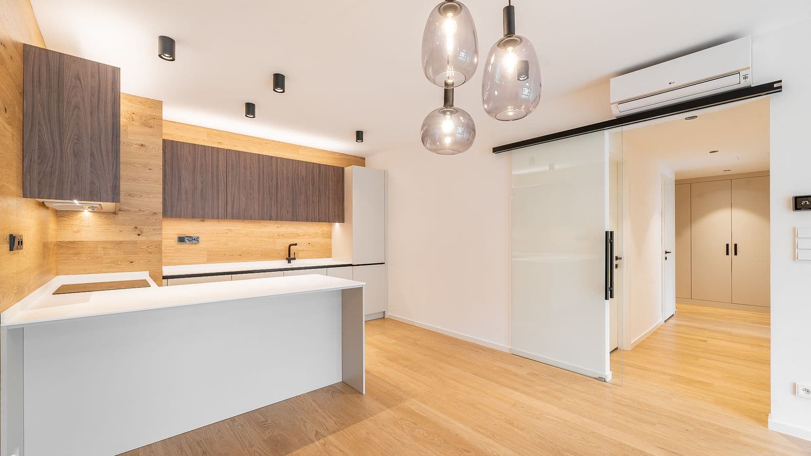 Moderní kuchyň s dřevěnou podlahou, dřevěným obkladem stěny, tmavě hnědými skříňkami, bílou pracovní deskou a třemi závěsnými světly.