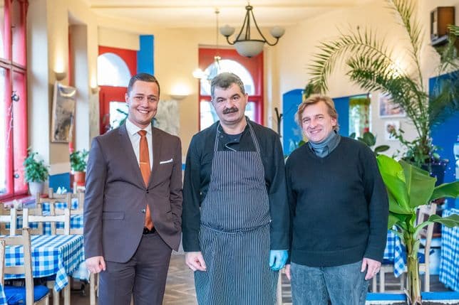 Three men standing in the interior of a Greek restaurant – on the left a man in a suit, in the center a chef in an apron, and on the right a man in a sweater. Blue and white tablecloths and plants in the background.