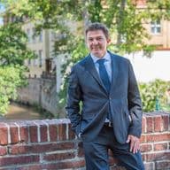 Muž v obleku opřený o cihlovou zeď s malebnou pražskou čtvrtí v pozadí, vedle protékající říčka a historická architektura Malé Strany