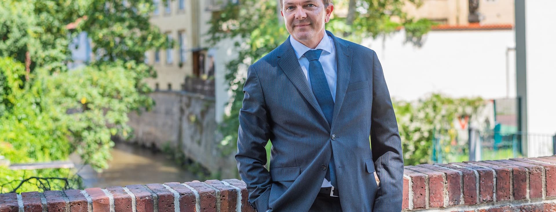 Muž v obleku opřený o cihlovou zeď s malebnou pražskou čtvrtí v pozadí, vedle protékající říčka a historická architektura Malé Strany