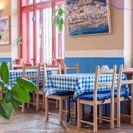 Interiér restaurace s modrobílými kostkovanými ubrusy, dřevěnými židlemi a okny s červenými rámy. Na stěnách visí fotografie řecké krajiny.