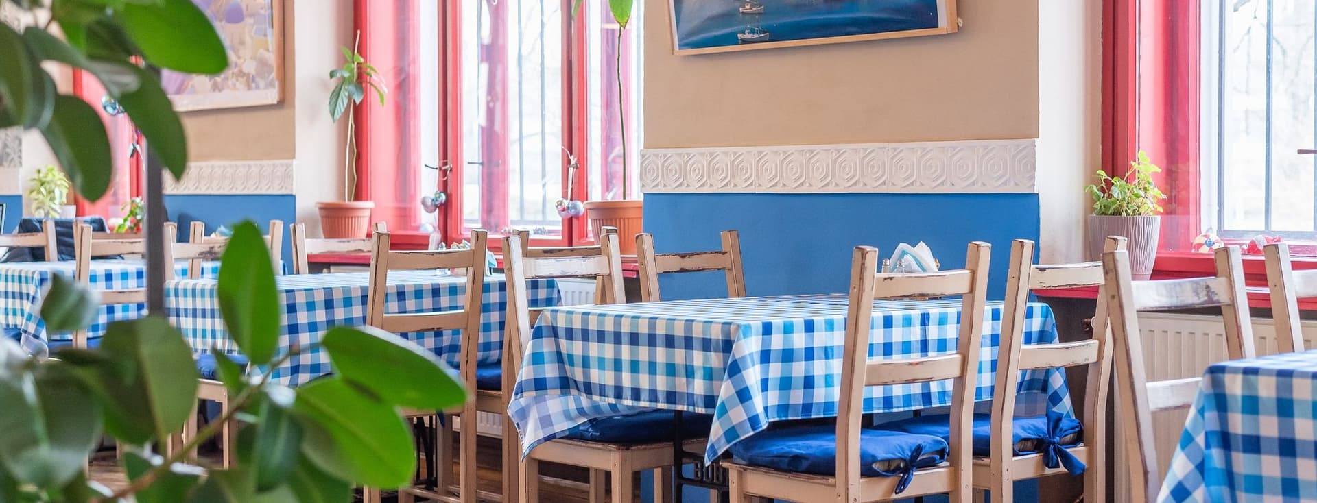 Interiér restaurace s modrobílými kostkovanými ubrusy, dřevěnými židlemi a okny s červenými rámy. Na stěnách visí fotografie řecké krajiny.