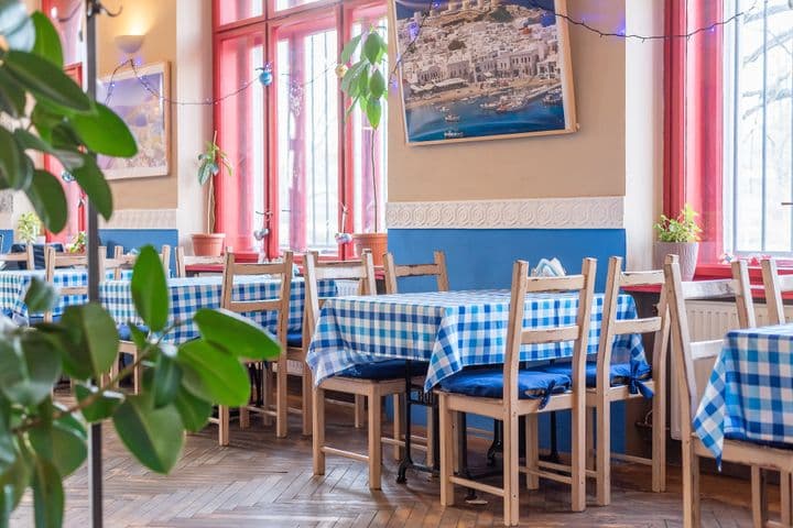 Interiér restaurace s modrobílými kostkovanými ubrusy, dřevěnými židlemi a okny s červenými rámy. Na stěnách visí fotografie řecké krajiny.