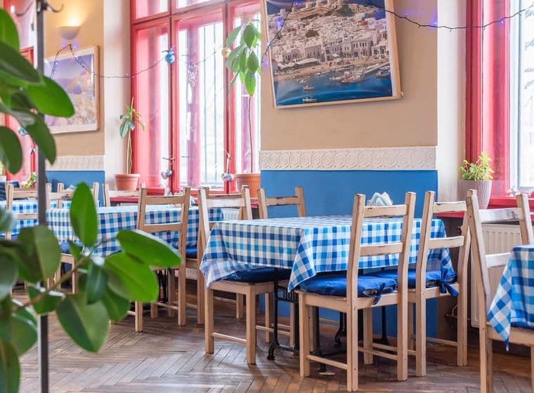 Interiér restaurace s modrobílými kostkovanými ubrusy, dřevěnými židlemi a okny s červenými rámy. Na stěnách visí fotografie řecké krajiny.