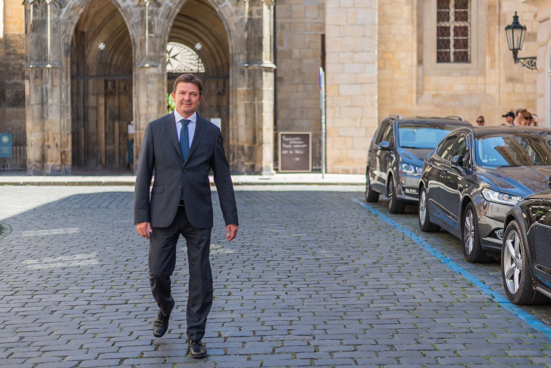 A man in a suit walks on a cobblestone street near parked cars, with a historic stone building