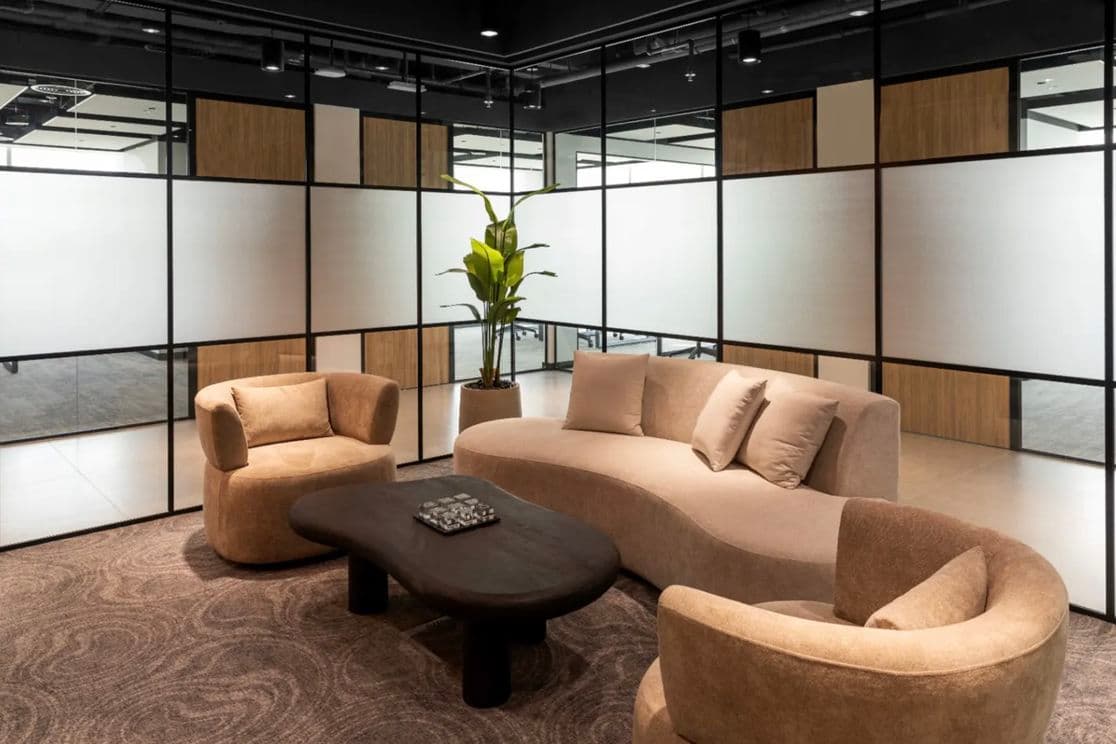 Modern office lounge with a curved beige sofa, two matching chairs, a dark wooden coffee table, and a potted plant against frosted glass dividers.