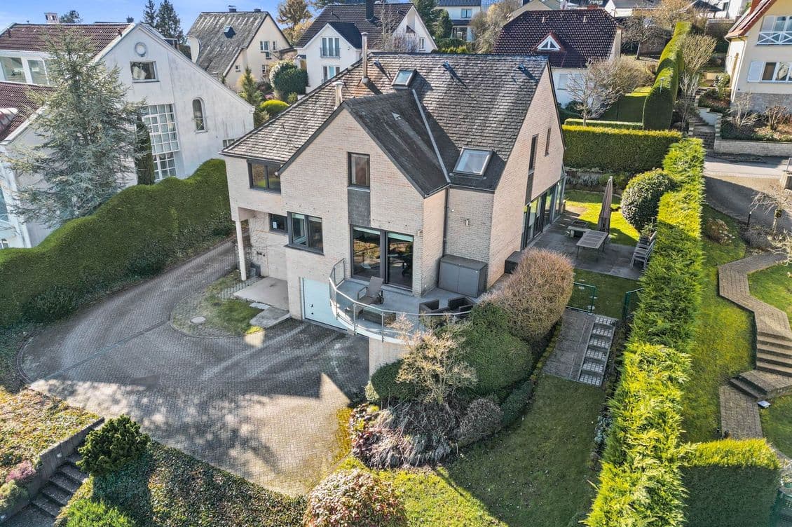 Aerial view of a modern two-story house with a sloped roof, surrounded by a landscaped yard, hedges, and neighboring homes in a suburban area.