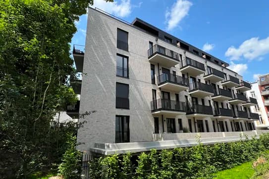 Modernes Wohnhaus aus grauen Ziegeln mit schwarzen Balkonen, umgeben von üppigem Grün unter blauem Himmel.