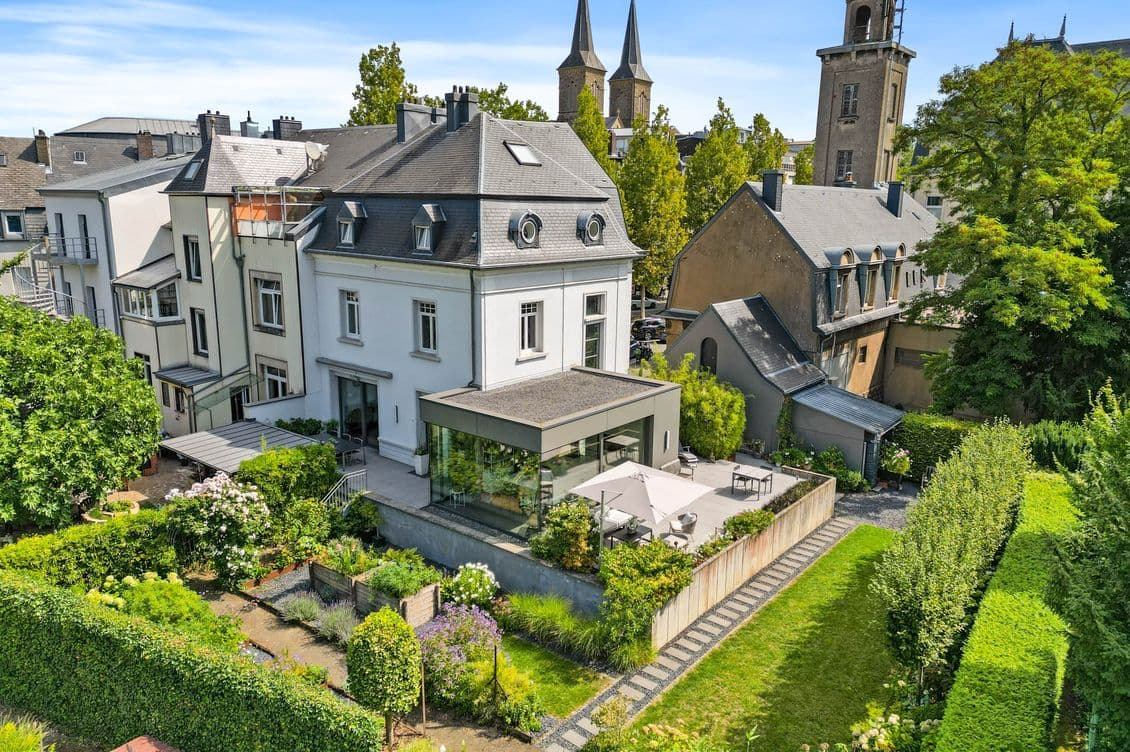 A sunlit European neighborhood with white and gray houses, a manicured garden, stone paths, and church towers in the distance.