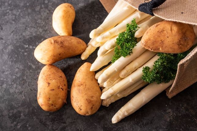 Potatoes and white asparagus spill from a burlap sack onto a dark textured surface, garnished with sprigs of parsley.