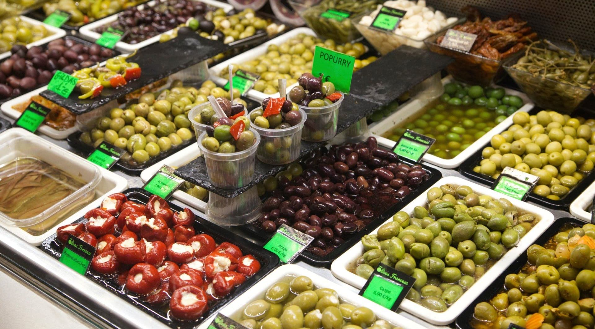 Nahaufnahme eines Markstandes mit verschiedenen Sorten grüner und schwarzer Oliven sowie gefüllten Peperoni