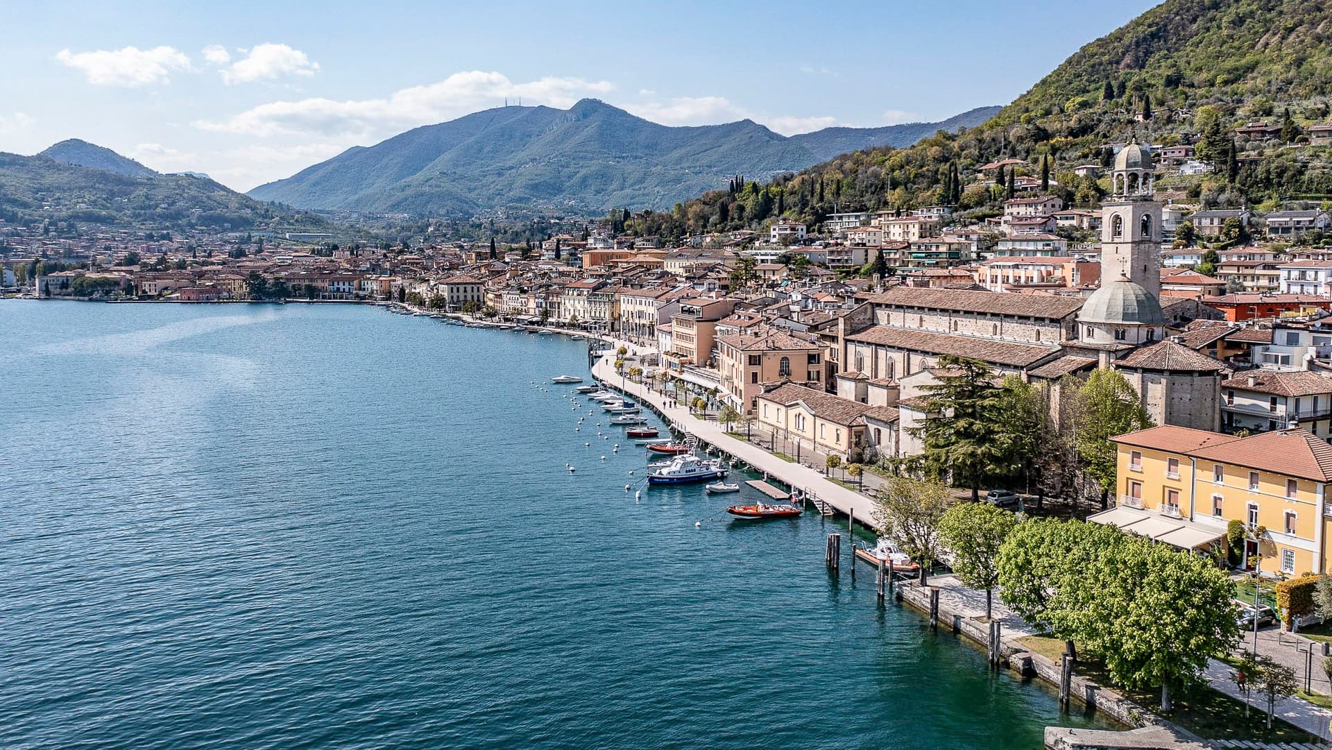 Veduta aerea di Salò sul Lago di Garda, con case colorate affacciate sulla riva, barche ormeggiate nel porto e le acque blu del lago sullo sfondo.