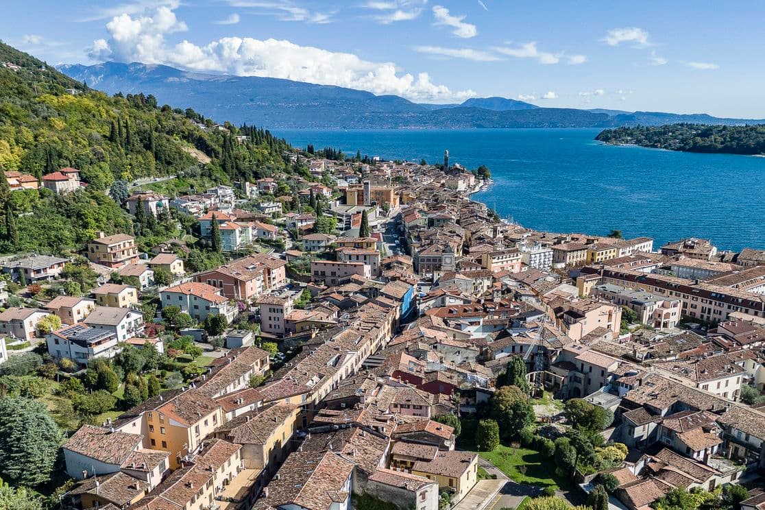 Veduta aerea di una pittoresca città lacustre con tetti in terracotta, circondata da una vegetazione lussureggiante e da un vasto lago blu sotto un cielo parzialmente nuvoloso.
