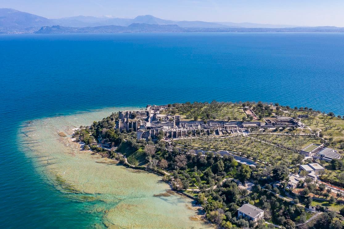 Luftaufnahme einer historischen Küstenstätte mit antiken Ruinen, umgeben von türkisfarbenem Wasser und üppigem Grün, mit Bergen im Hintergrund.