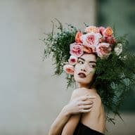 Woman with a floral headpiece of pink and orange roses, surrounded by greenery, poses against a blurred background.