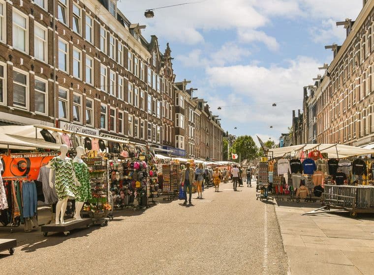 Street view of Albert Cuyp Market in Amsterdam, Netherlands. Vendors sell clothing and souvenirs from stalls. People walk along the street.