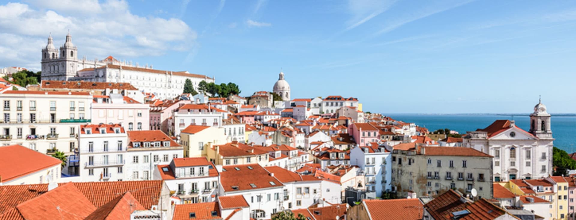Lisbon cityscape with red-tiled roofs and white buildings under a blue sky with contrails.