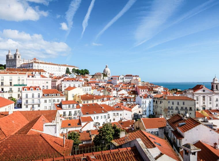 Lisbon cityscape with red-tiled roofs and white buildings under a blue sky with contrails.