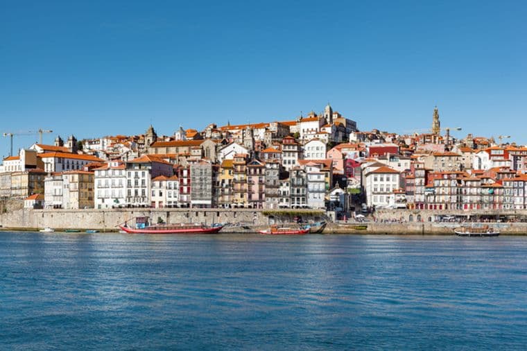 Skyline-Ansicht von Porto, Portugal, mit farbenfrohen Gebäuden und rot gedeckten Häusern auf einem Hügel mit Blick auf den Douro. Boote liegen entlang der Uferpromenade vor Anker.
