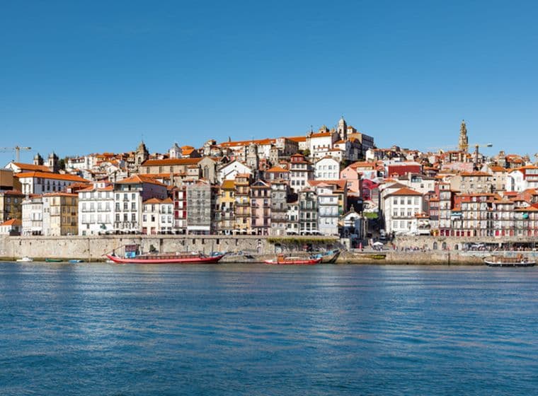 Vista panoràmica de Porto, Portugal, amb edificis colorits i cases amb teulades vermelles en un turó amb vistes al riu Douro. Hi ha vaixells atracats al llarg del passeig marítim.