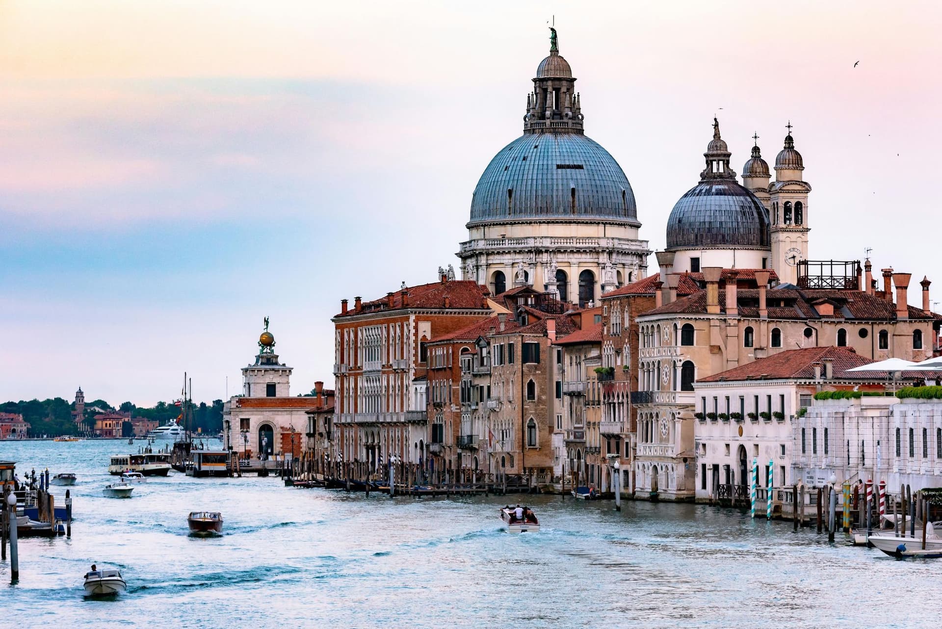 Venice canal scene with historic buildings, including a domed church, boats on the water, and a soft pink and blue sky at sunset.