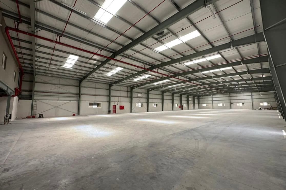 Spacious, empty warehouse interior with a high ceiling, metal beams, white walls, and a polished concrete floor, illuminated by overhead lights.