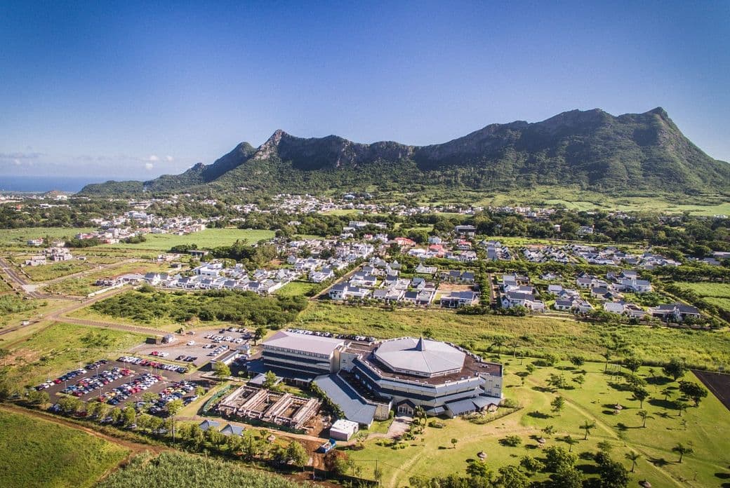 Aerial view of Moka with a large building in the foreground, surrounded by greenery and mountains under a clear blue sky.