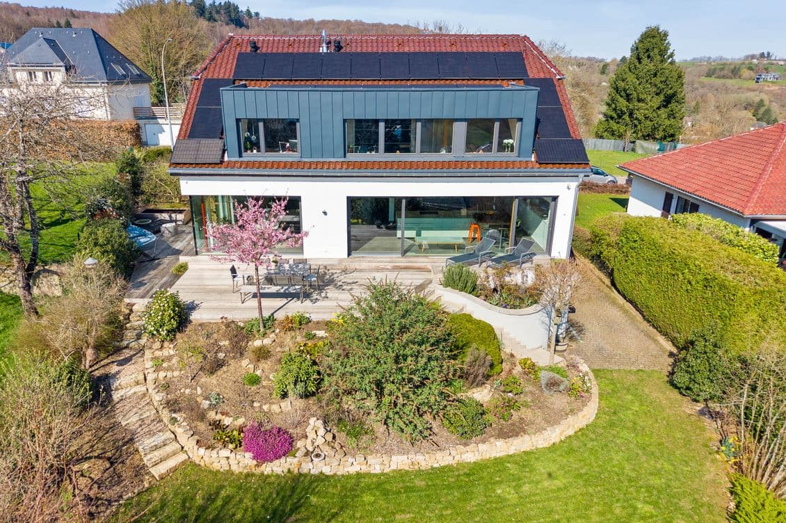 An aerial view of a modern house with a red-tiled roof, solar panels, large glass windows, and a terraced backyard with stone landscaping.