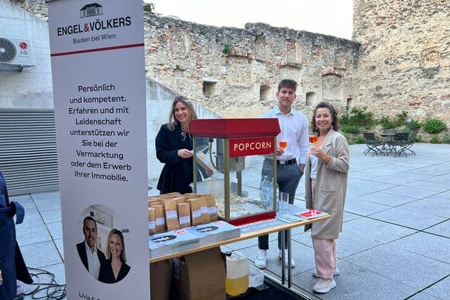 Immobilienmakler-Team von Engel & Völkers mit Popcorn-Stand vor historischer Steinmauer, Banner mit Unternehmensphilosophie im Vordergrund
