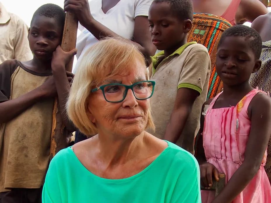 A white woman wearing turquoise glasses stands among a group of African children and adults.