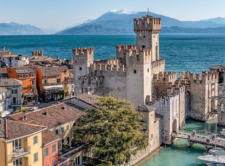 Malerischer Blick auf ein mittelalterliches Schloss an einem See mit Bergen im Hintergrund, umgeben von farbenfrohen Gebäuden und in der Nähe vertäuten Booten.