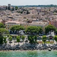 Luftansicht einer italienischen Küstenstadt mit historischen Gebäuden, baumumsäumter Promenade und felsiger Küstenlinie entlang des grünen Meeres.